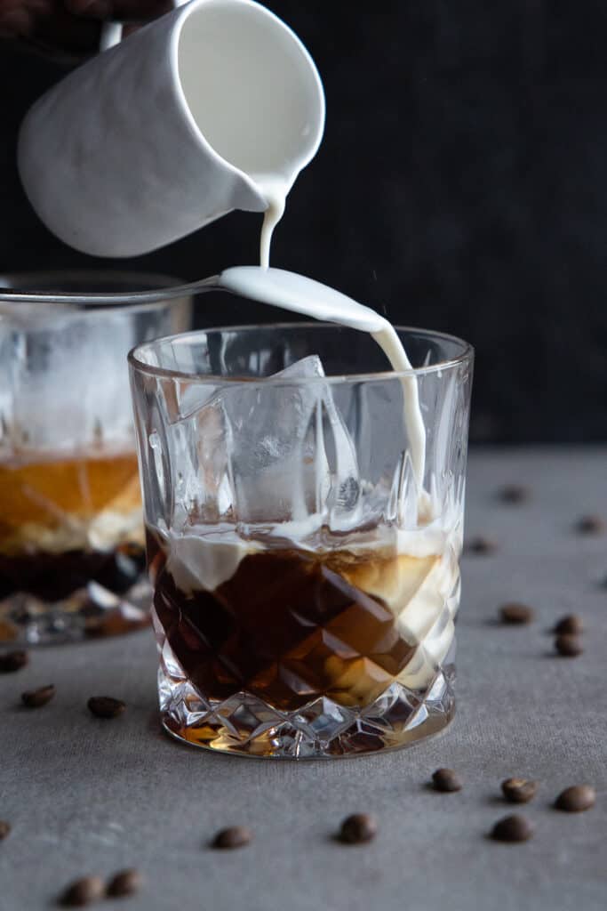 Pouring cashew milk over the back of a spoon into glass to create a vegan white Russian.